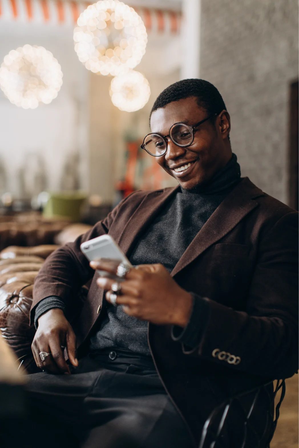 A businessman in glasses and a brown suit, sitting down and smiling while looking at his cell phone