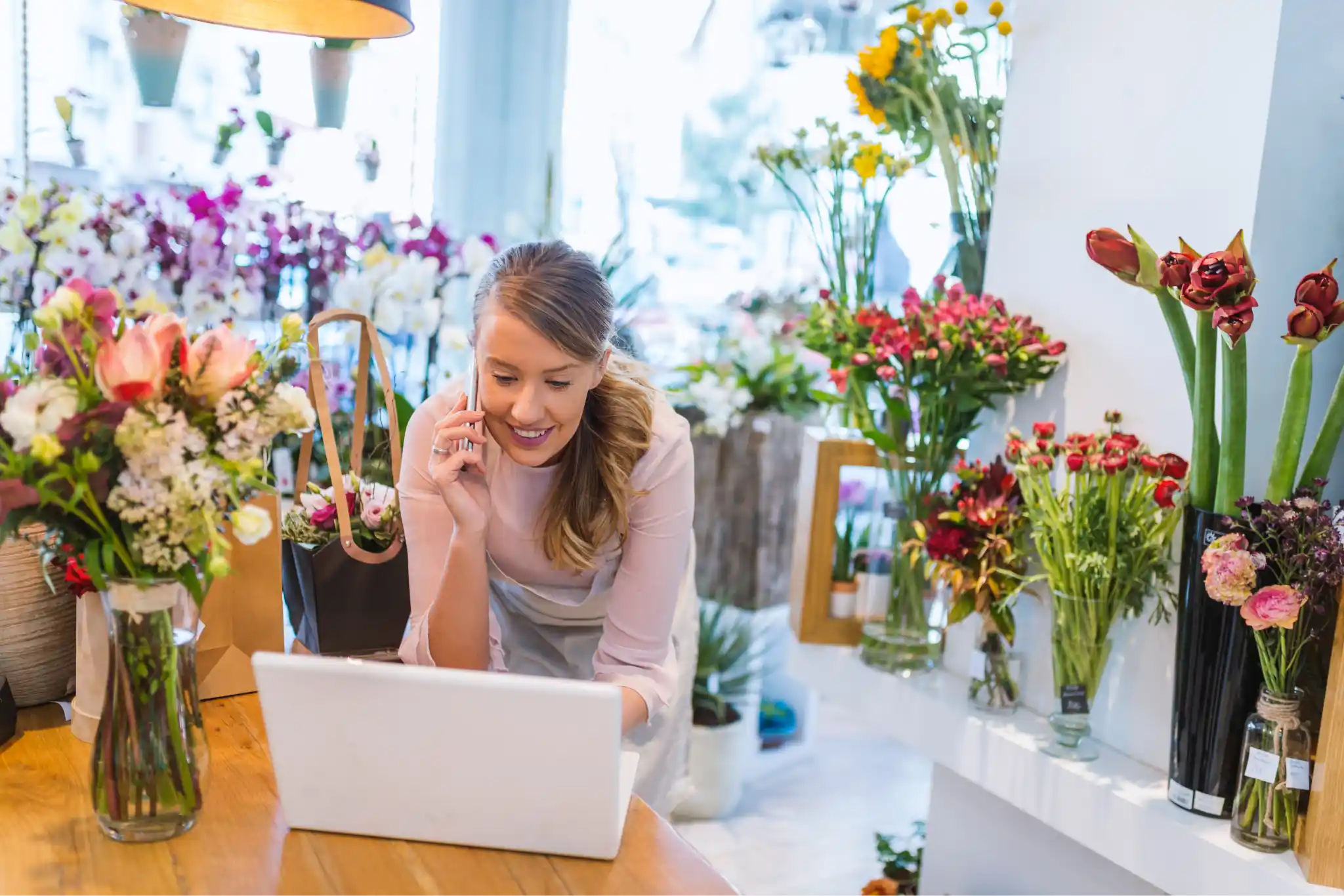 A florist talking on the phone and using a laptop while surrounded by flowers.