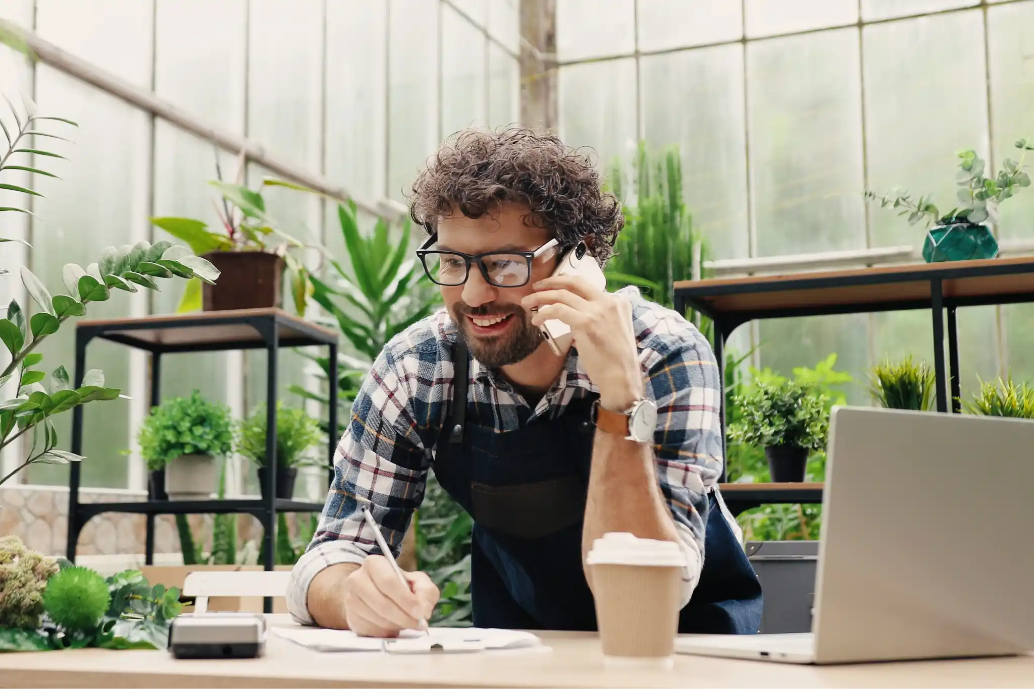 Man in an apron in a plant nursery, talking on a phone, holding a pen, and a laptop on the counter