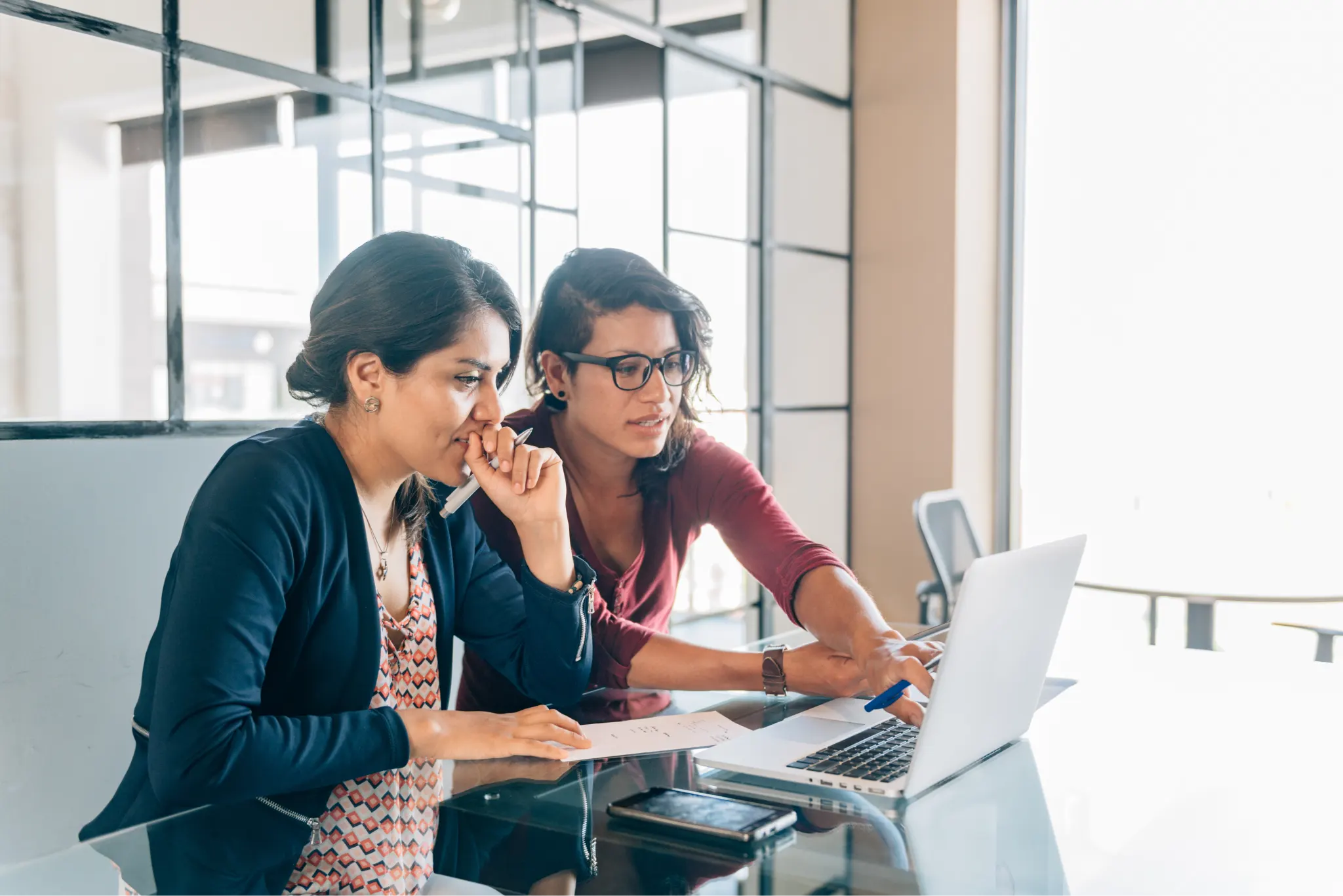 Two businesswomen sitting a table, focused on a laptop screen.